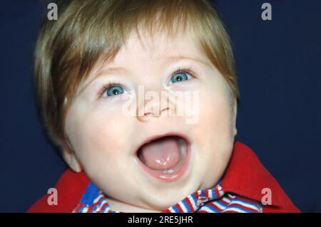 Das Baby hat den Mund weit geöffnet und seine Augen funkeln. Die Nahaufnahme seines Gesichts zeigt zwei kleine Zähne in seinem offenen Mund. Stockfoto