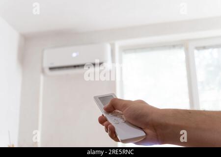 Fernbedienung mit der Hand, die auf die Klimaanlage gerichtet ist. Junger Mann, der die Klimaanlage mit Fernbedienung anmacht. Erhöhte Temperaturen, heißes Wetter. Stockfoto