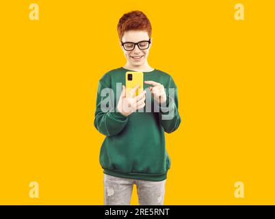 Ein hübscher lächelnder Junge mit Brille und rotem Haar, der auf seinem Mobiltelefon durch die Speisekarte blättert oder ein Foto macht. Stockfoto