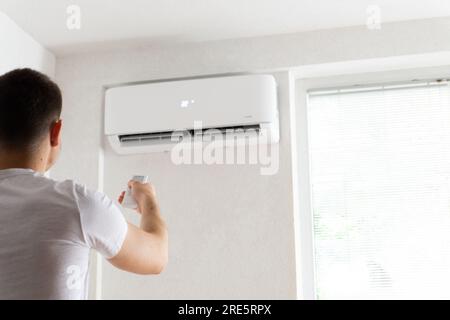 Der junge Mann schaltet die Klimaanlage ein. Hübscher junger Mann, der die Klimaanlage mit Fernbedienung anmacht. Steigende Temperaturen, heißes Wetter. Stockfoto