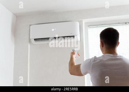 Der junge Mann schaltet die Klimaanlage ein. Hübscher junger Mann, der die Klimaanlage mit Fernbedienung anmacht. Steigende Temperaturen, heißes Wetter. Stockfoto