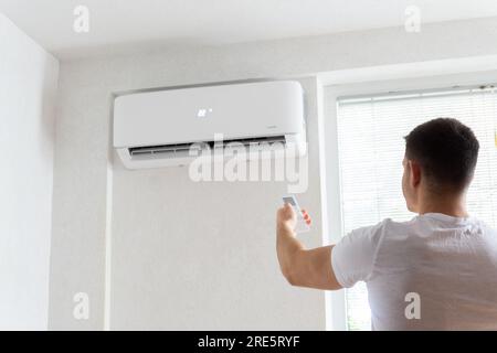 Der junge Mann schaltet die Klimaanlage ein. Hübscher junger Mann, der die Klimaanlage mit Fernbedienung anmacht. Steigende Temperaturen, heißes Wetter. Stockfoto