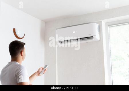 Der junge Mann schaltet die Klimaanlage ein. Hübscher junger Mann, der die Klimaanlage mit Fernbedienung anmacht. Steigende Temperaturen, heißes Wetter. Stockfoto