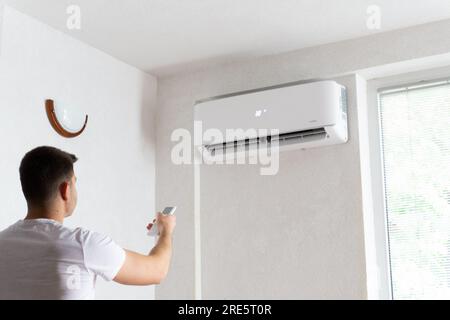 Der junge Mann schaltet die Klimaanlage ein. Hübscher junger Mann, der die Klimaanlage mit Fernbedienung anmacht. Steigende Temperaturen, heißes Wetter. Stockfoto