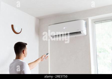 Der junge Mann schaltet die Klimaanlage ein. Hübscher junger Mann, der die Klimaanlage mit Fernbedienung anmacht. Steigende Temperaturen, heißes Wetter. Stockfoto