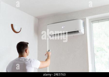 Der junge Mann schaltet die Klimaanlage ein. Hübscher junger Mann, der die Klimaanlage mit Fernbedienung anmacht. Steigende Temperaturen, heißes Wetter. Stockfoto