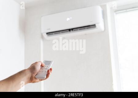 Fernbedienung mit der Hand, die auf die Klimaanlage gerichtet ist. Junger Mann, der die Klimaanlage mit Fernbedienung anmacht. Erhöhte Temperaturen, heißes Wetter. Stockfoto