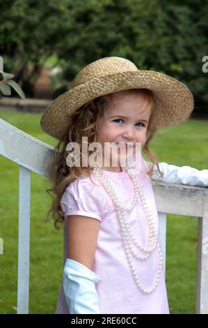 Ein hübsches kleines Mädchen, das einen rosa Hut und ein Kleid trägt, legt eine Hand mit Handschuhen auf ein rustikales Holztor. Sie trägt auch eine lange Birnensträhne. Stockfoto