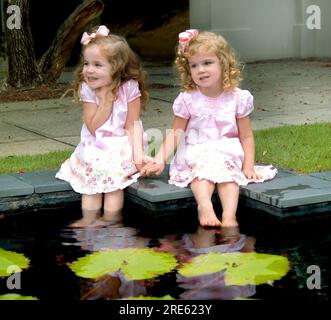 Zwei hübsche kleine Mädchen sitzen nebeneinander am Rand eines lilly-Pad-Teiches. Sie machen sich die Füße nass und lachen und lächeln. Die Kleider sind rosa. Stockfoto