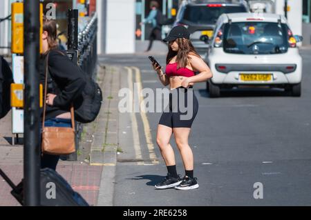 Junge Frau, die für den Sommer gekleidet ist und auf einer Straße steht, mit einem Mobiltelefon, Smartphone oder Mobiltelefon in England, Großbritannien. Stockfoto