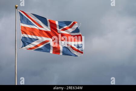 Union Jack, Flagge des Vereinigten Königreichs Großbritannien und Nordirland, fliegt im starken Wind auf einem Stab gegen bewölkten Himmel. Stockfoto