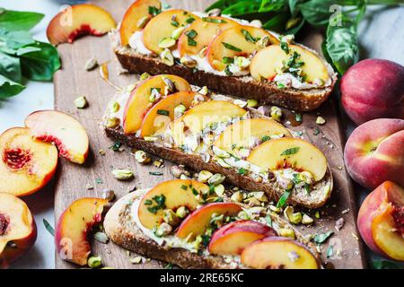 Frischer Vollkorntoast mit Pfirsich und Pistazien, mit ausgeschlagenem veganem Ricotta-Käse, Basilikum und Honig. Selektiver Fokus mit verschwommenem Vorder- und Hintergrund Stockfoto