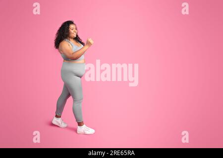 Frohe schwarze, übergroße Frau in Sportbekleidung, die Jogging und Workout genießt, pinkfarbener Hintergrund, volle Länge, freier Platz Stockfoto