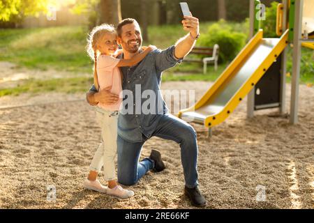 Spielplatzerinnerungen. Dad und ihre hübsche Tochter machen Selfie auf dem Spielplatz im Park, Mädchen umarmt ihren Vater Stockfoto