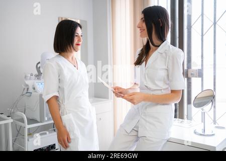 Zwei Ärztinnen stehen in der medizinischen Klinik und diskutieren die Ergebnisse der Patientenuntersuchungen. Sie sehen sich an und lächeln. Spezialisten im Gesundheitswesen führen ein professionelles Gespräch. Stockfoto