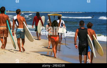 Rückansicht einer Gruppe von Surfern, die den Strand entlang laufen und Surfbretter am Ufer des Gilgo Beach mit sich führen. Stockfoto