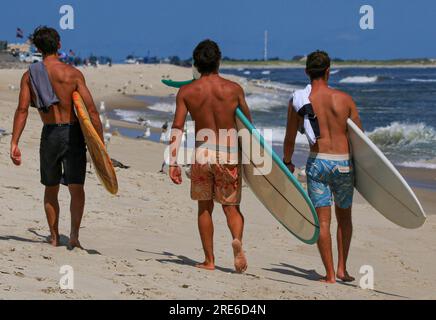 Rückansicht von drei Jungen, die am Strand spazieren und am Ufer von Gilgo Beach Long Island Surfboads tragen. Stockfoto