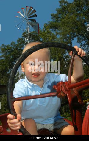 Das Kleinkind nimmt das Rad eines Traktors. Er will lernen, wie man Landwirtschaft treibt. Seine Hände greifen das Lenkrad. Im Hintergrund steht eine Windmühle. Stockfoto