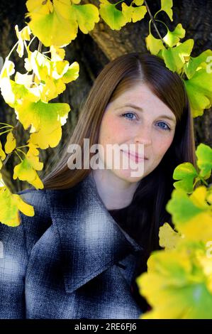 Eine wunderschöne junge Frau lehnt sich an einem kühlen Herbstnachmittag in Memphis, Tennessee, gegen einen Ghinko-Baum. Gelbe Blätter umrahmen Nahaufnahmen ihres Gesichts. Stockfoto