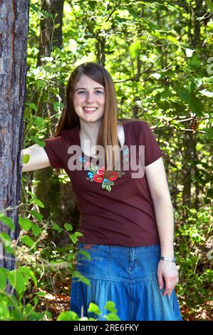 Glückliche junge Frau lehnt sich im Wald an einen Baum. Sie trägt einen Jeansrock und ein braunes Hemd. Ihr Lächeln ist voller Glück und Frieden. Stockfoto