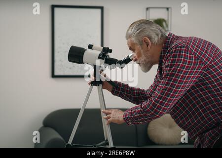 Seniorenmann, der Sterne durch ein Teleskop im Zimmer sieht Stockfoto