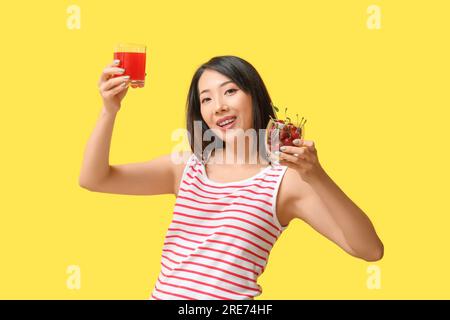 Wunderschöne asiatische Frau mit Kirschen und einem Glas Saft auf gelbem Hintergrund Stockfoto