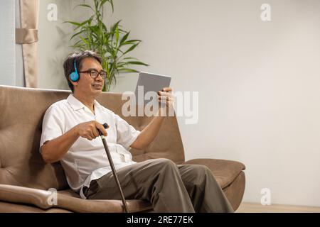 Sarkopenie älterer Mann benutzt ein digitales Tablet und Kopfhörer auf dem Sofa, um sich im Pflegeheim zu entspannen. Stockfoto