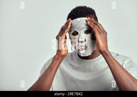 Studioporträt eines jungen, lächelnden schwarzen Mannes, der eine feuchtigkeitsspendende Papiermaske auf sein Gesicht aufträgt. Glücklicher afroamerikanischer Millennialmensch verwendet Schönheitsprodukte in Stockfoto