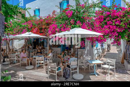 Gäste genießen Getränke und Frühstück in einem traditionellen Straßencafé in einer kopfsteingepflasterten engen Gasse mit Bougainvillea-Baum in Antiparos, Griechenland. Stockfoto