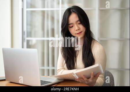 Eine wunderschöne junge asiatische Studentin konzentriert sich darauf, Informationen in einem Buch zu lesen, während sie zu Hause online lernt. Bildungs- und Menschenkonzept Stockfoto