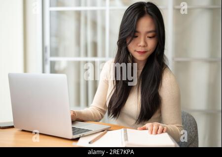 Eine wunderschöne junge asiatische Studentin konzentriert sich darauf, Informationen in einem Buch zu lesen, während sie zu Hause online lernt. Bildungskonzept Stockfoto