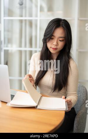 Das Porträt einer wunderschönen jungen asiatischen Studentin konzentriert sich darauf, Informationen in einem Buch zu lesen, während sie zu Hause online studiert. Stockfoto