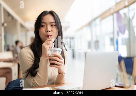 Eine glückliche und charmante junge asiatische Frau, die ihren Eiskaffee genießt, während sie aus der Ferne in einem Café arbeitet. Getränke- und Lifestyle-Konzept Stockfoto