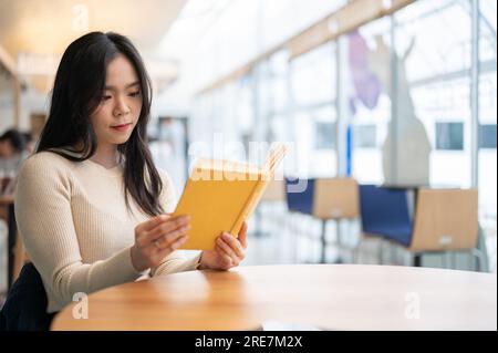 Eine wunderschöne junge Asiatin liest ein Buch, während sie sich an ihrem Wochenende in einem Café entspannen. college-Student, Freiberufler, Lifestyle Stockfoto