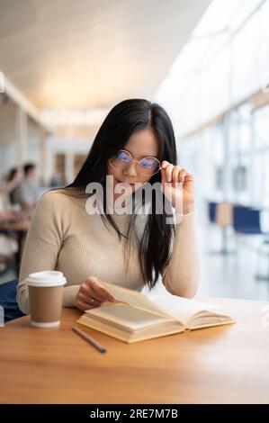 Das Porträt einer schönen, klugen, jungen asiatischen Studentin mit Brille konzentriert sich darauf, ein Buch zu lesen, während sie in einem Café in einem Co-Working-Bereich sitzt. Stockfoto