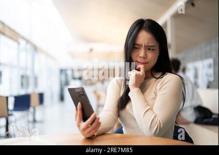 Eine unsichere und umsichtige junge Asiatin sieht auf ihren Smartphone-Bildschirm mit einem ernsthaften und zweifelhaften Gesicht, während sie in einem Café sitzt. Ich mache mir Sorgen Stockfoto