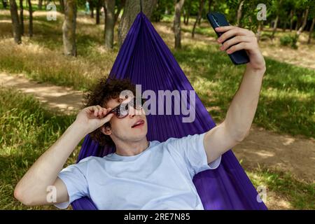 Ein junger Mann macht ein Selfie am Telefon und entspannt sich in einer lila Hängematte in der Natur, genießt die Stille. Stockfoto