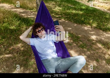 Ein junger Mann macht ein Selfie am Telefon und entspannt sich in einer lila Hängematte in der Natur, genießt die Stille. Stockfoto