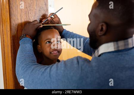 Fröhlicher afroamerikanischer Vater, der zu Hause seinen Sohn misst und die Höhe am Türrahmen markiert Stockfoto