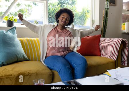 Eine glückliche afroamerikanerin in Übergröße, die auf dem Sofa sitzt und sich zu Hause streckt Stockfoto