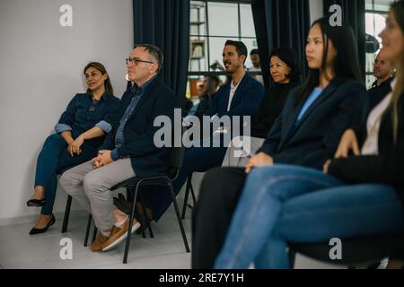 Gruppe von Multiethnen in formeller Kleidung, die an einem modernen Arbeitsplatz auf Stühlen sitzen und während des Seminars aufmerksam wegschauen Stockfoto