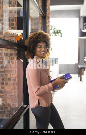 Porträt einer glücklichen, birassistisch-lässigen Geschäftsfrau, die ein Smartphone benutzt und Dateien im Büro hält Stockfoto