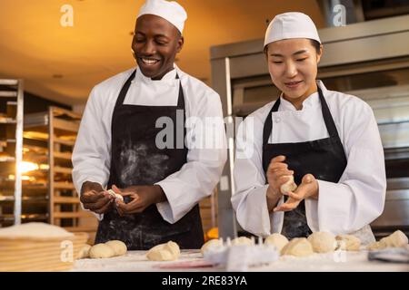 Glückliche, vielfältige Bäcker, die Schürzen in der Bäckerei tragen und Teigrollen machen Stockfoto