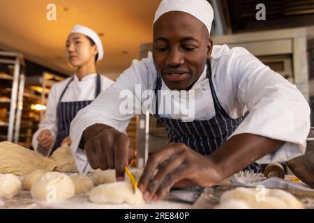 Glückliche, vielfältige Bäcker, die Schürzen in der Bäckerei tragen und Teigrollen machen Stockfoto