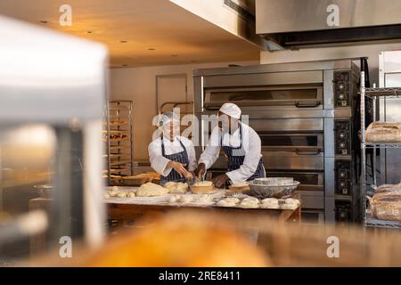 Glückliche, vielfältige Bäcker, die Schürzen in der Backküche tragen und Teig schneiden Stockfoto