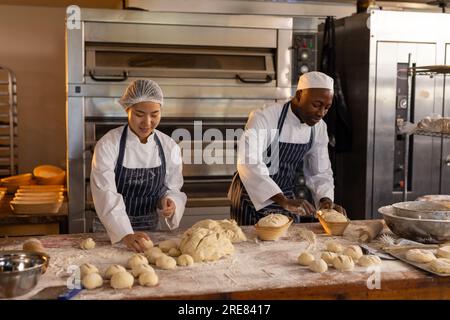 Glückliche, vielfältige Bäcker, die Schürzen in der Bäckerei tragen und Teigrollen machen Stockfoto