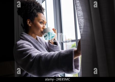 Eine birassische Frau im Bademantel, die Vorhänge öffnet und Kaffee trinkt Stockfoto