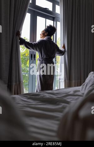 Der Rücken einer glücklichen, birassischen Frau im Bademantel, die die Vorhänge im Schlafzimmer öffnet Stockfoto