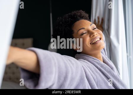 Glückliche, birassische Frau im Bademantel, die die Vorhänge im Schlafzimmer öffnet Stockfoto