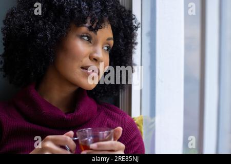 Rücksichtsvolle, birassische Frau, die am Fenster sitzt und zu Hause Tee trinkt Stockfoto